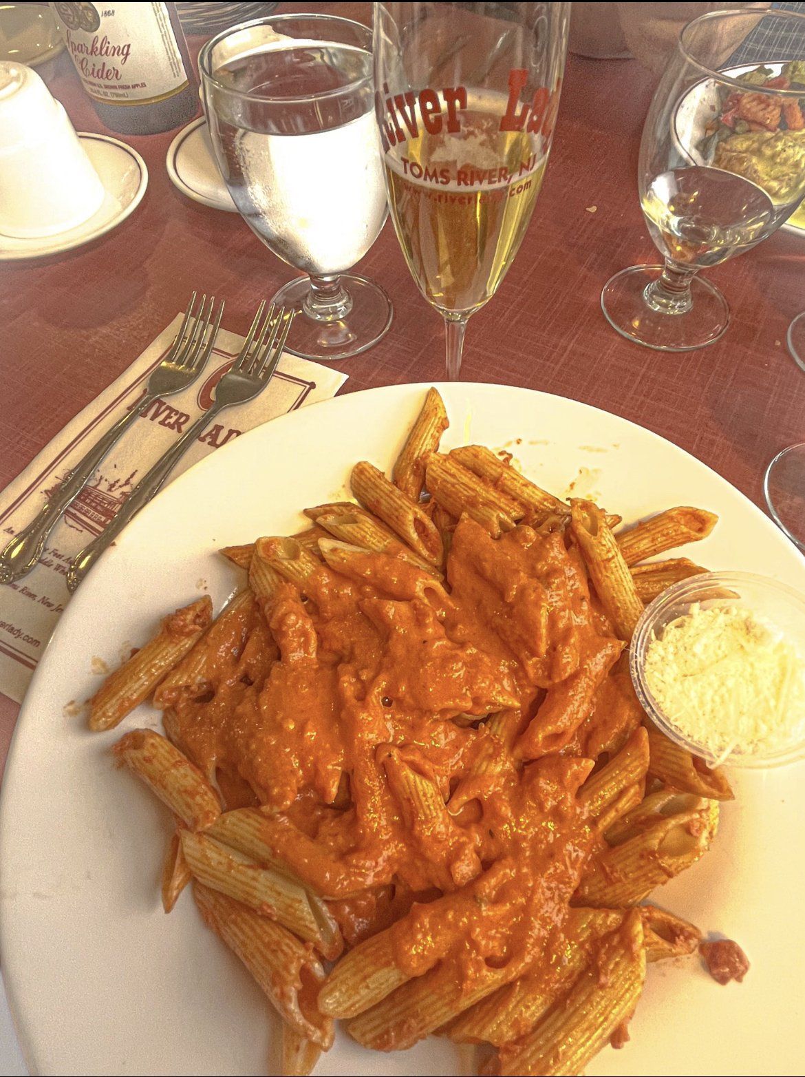 Penne pasta with tomato cream sauce and grated cheese, on a white plate at a restaurant table with drinks.