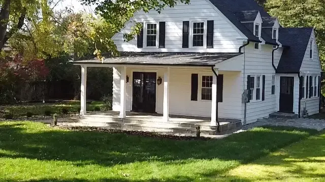A white house with a black roof and a porch surrounded by trees.