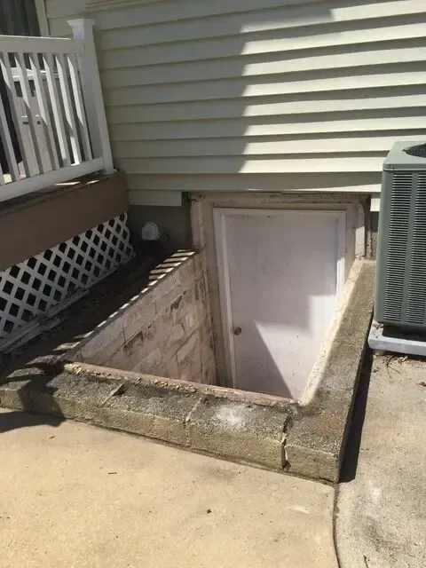 A basement door is built into the side of a house next to an air conditioner.