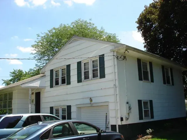 Two cars are parked in front of a white house with black shutters