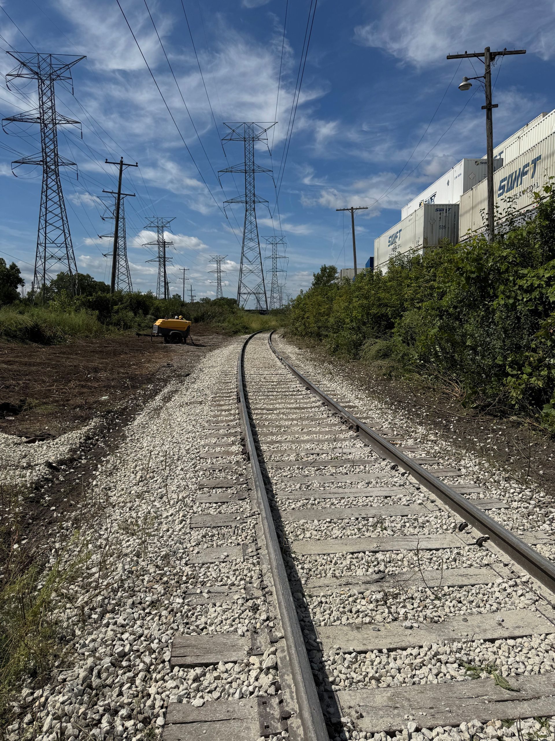 Railroad tracks curving through a gravel bed, passing power lines and a graffiti-covered wall under a blue sky.