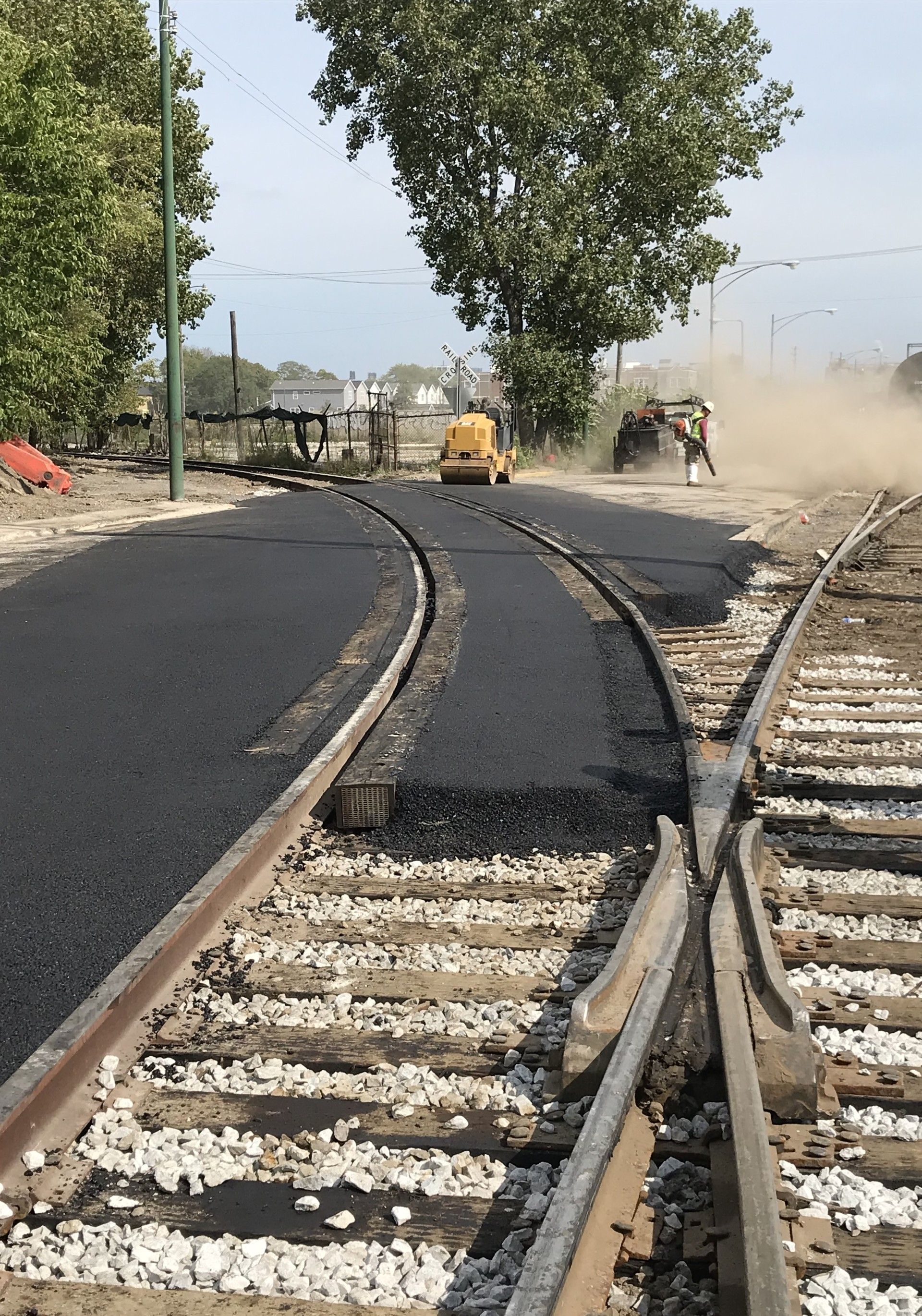 Asphalt being laid between railroad tracks. A small yellow machine and workers are present.