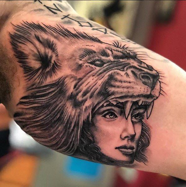 Tattoo of a woman's face with a lion's head above her. Black and white. On a man's arm.