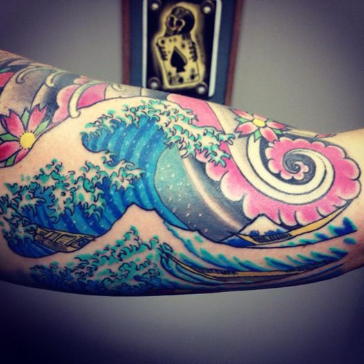 Colorful Japanese-style wave tattoo on an arm with pink flowers, a boat, and mountains.