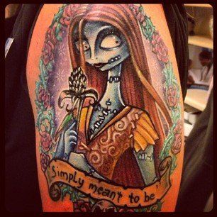 Tattoo of Sally from The Nightmare Before Christmas, framed by flowers, with a banner: