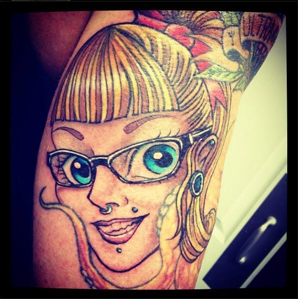 Tattoo of a woman with blonde hair, glasses, and piercings. Smiling, with blue eyes.