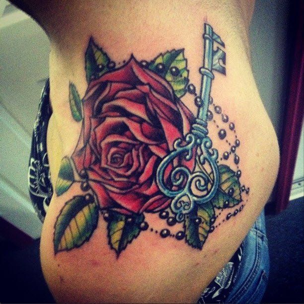 Tattoo of a red rose, ornate blue key, leaves, and pearl beads on a person's side.
