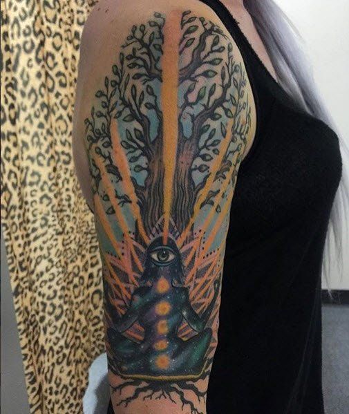 Tattoo of a meditating figure with open third eye under a tree, with chakra colors and rays of light.