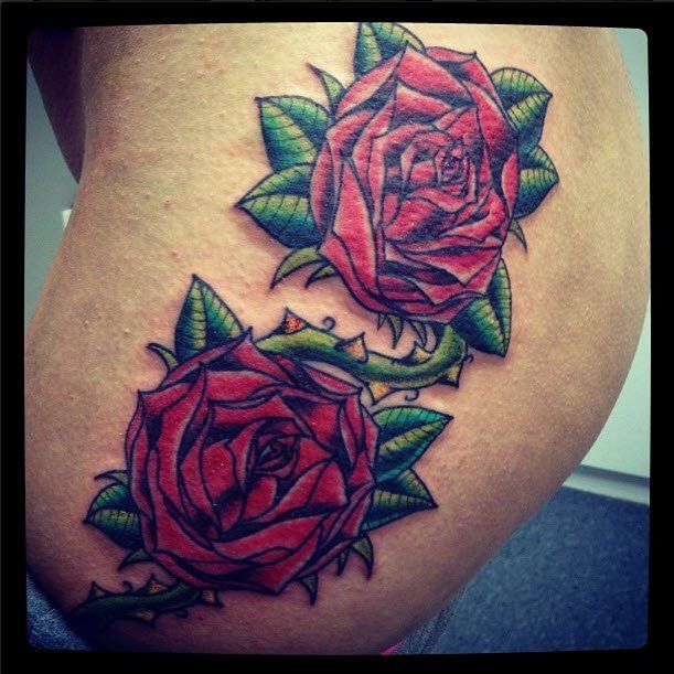 Tattoo of two red roses with green leaves and thorny vines on skin.