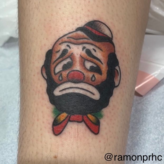 Traditional tattoo of a sad clown with a red nose, hat, and bow tie, on a leg.