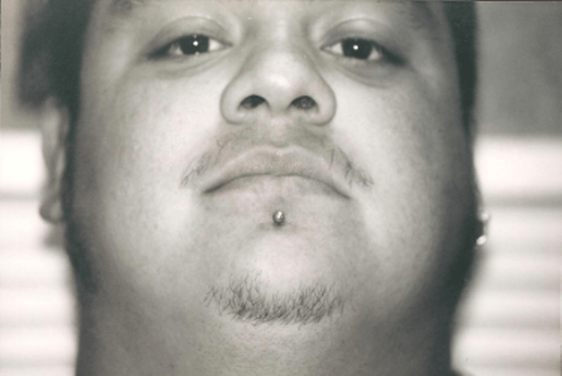 Close-up of a man with a goatee, chin piercing, and earring, looking up at the camera.