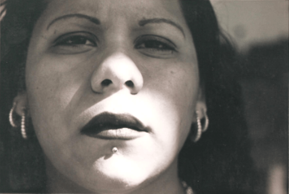Close-up of a woman with dark lipstick, eyebrow piercings, and earrings, looking directly at the camera.