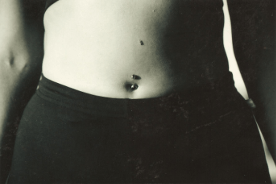 Person's torso with a belly button piercing; wearing black pants. Black and white photo.