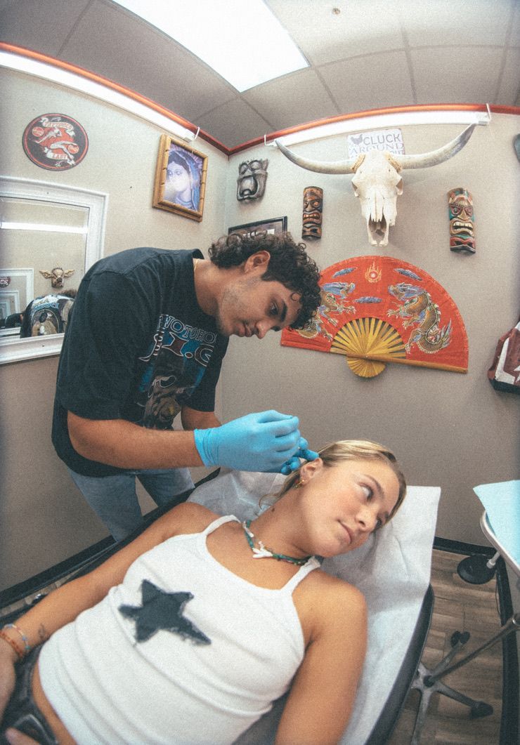 Tattoo artist piercing woman's ear in a studio. Woman lies on a bed, man wears gloves. Skull and art on wall.