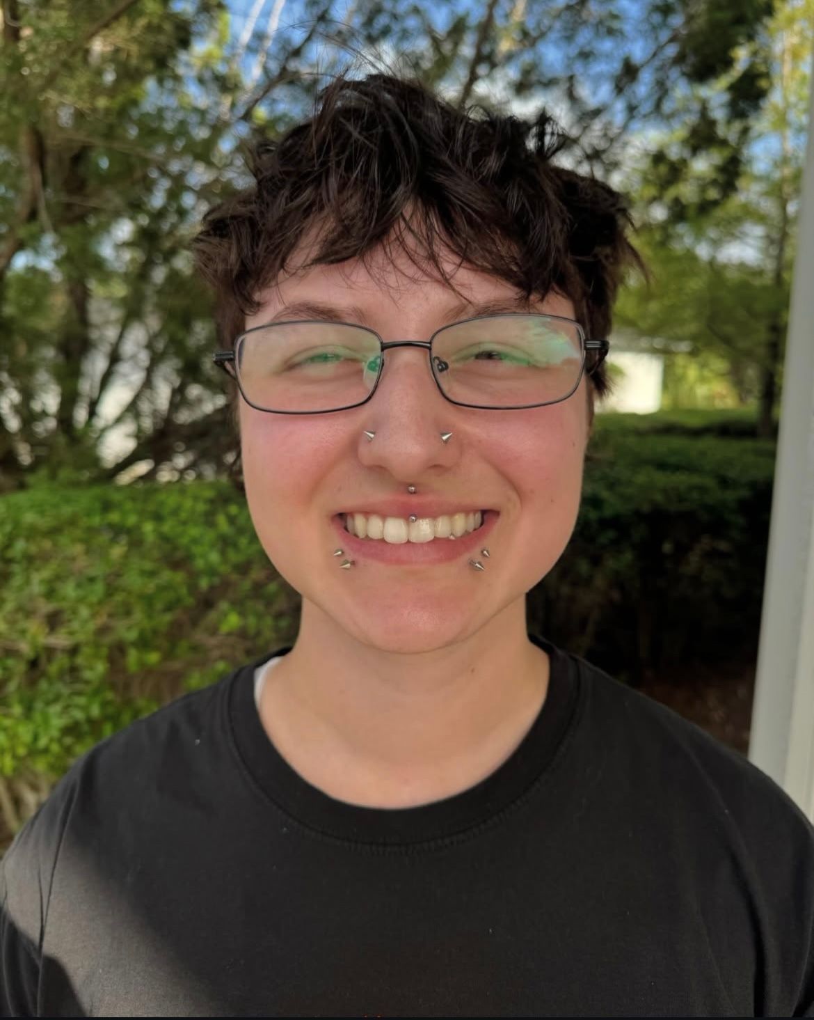Person with short, dark hair smiles, wearing glasses and facial piercings outdoors in front of greenery.