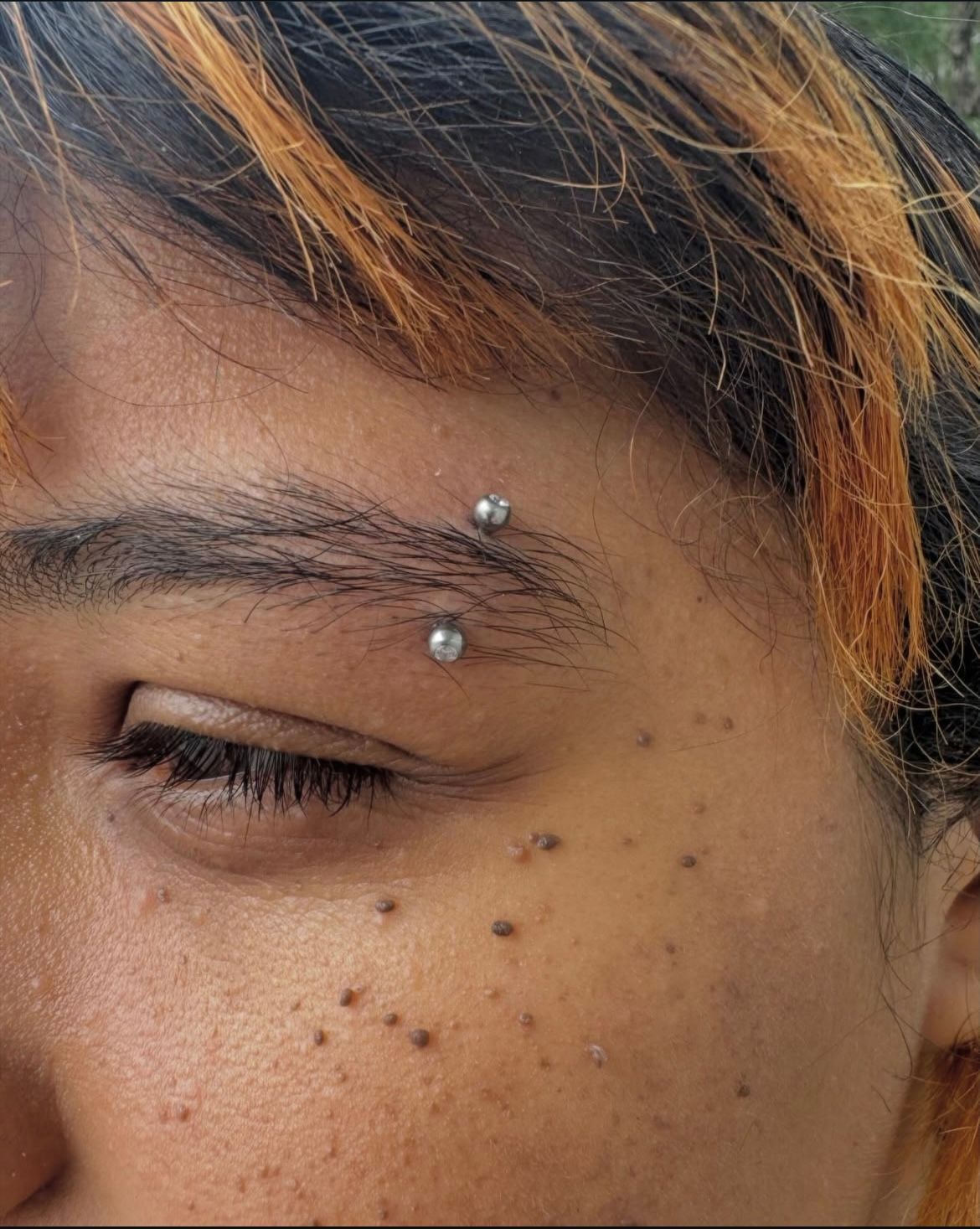 Close-up of person with eyebrow piercing, freckles, and highlighted hair.