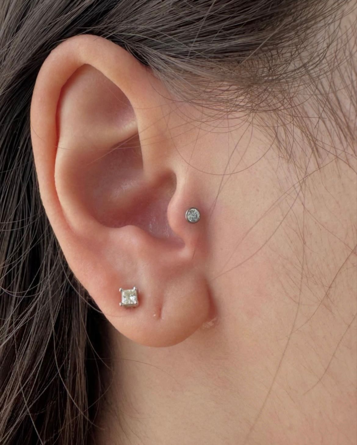 Close-up of ear with two small, sparkling earrings; one square, one round.