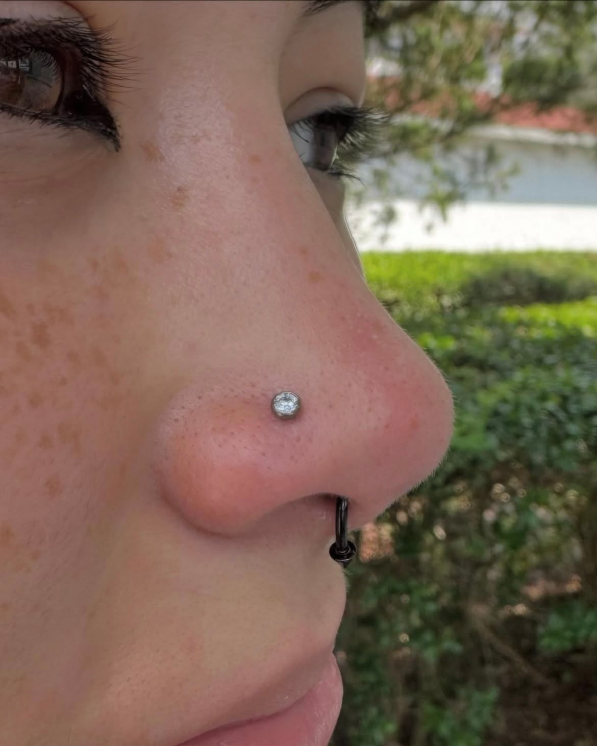 Close-up of a person's nose with a nostril piercing and a septum ring, outdoors.