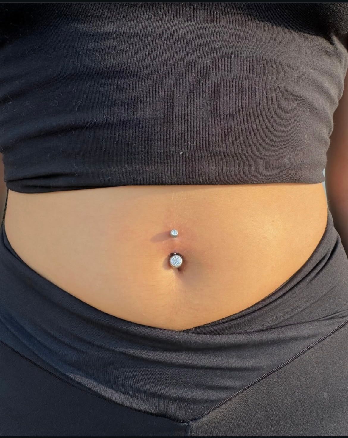 Person's belly with a belly button piercing. Wearing black top and pants. Sunny outdoor setting.
