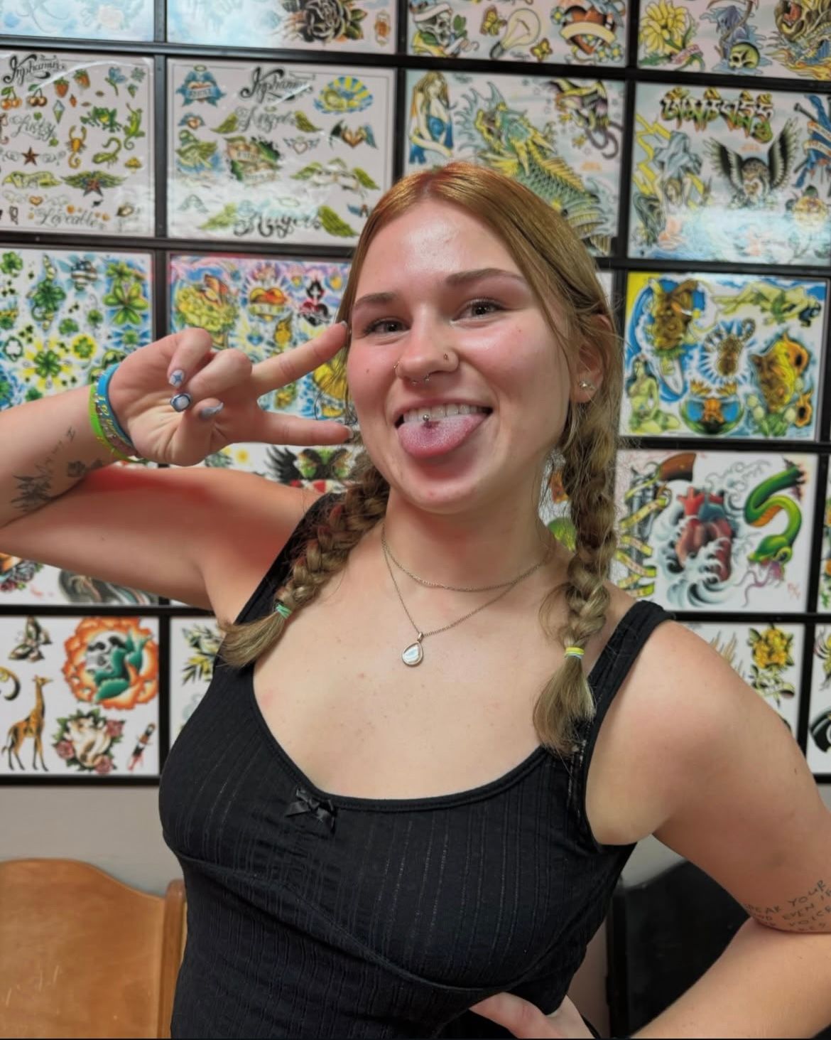 Young woman with tongue piercing, braids, and peace sign, in front of tattoo flash art.