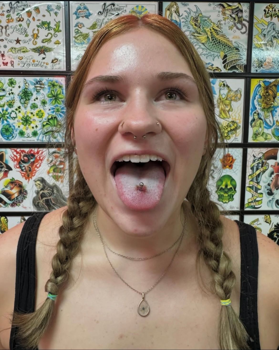 Woman with pigtails, open mouth, tongue piercing, and nose piercings, in a tattoo shop, looking up.