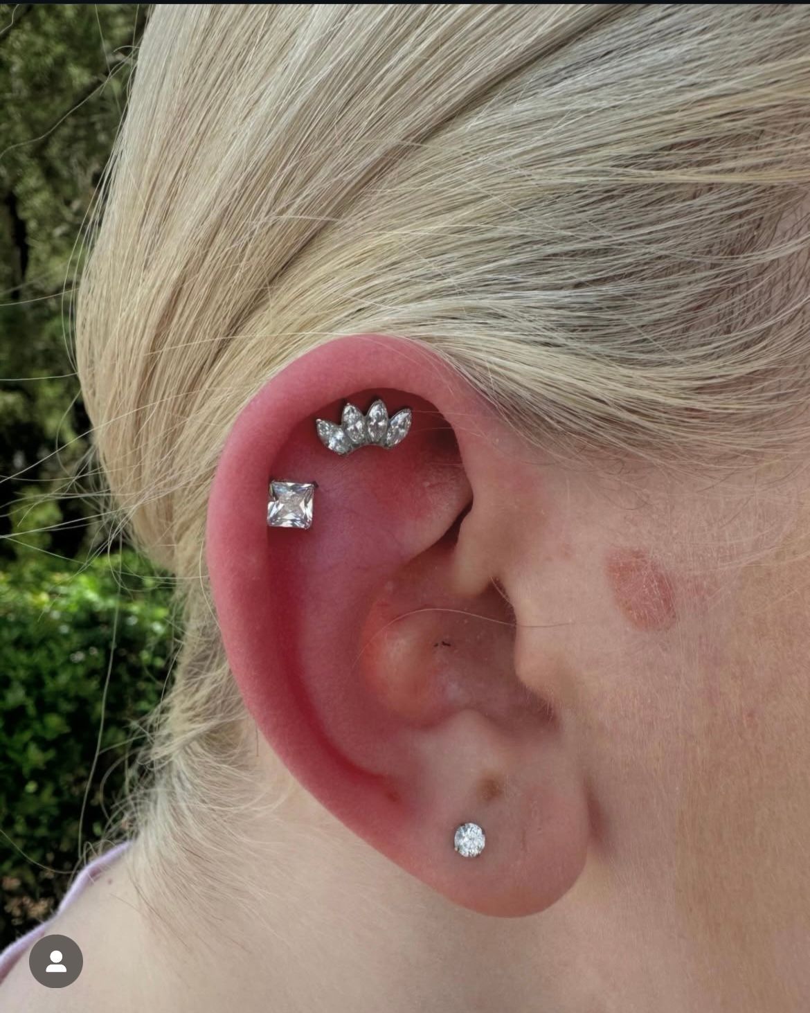 Ear with multiple piercings: helix, conch, and lobe, all with silver-toned gems. Blonde hair.