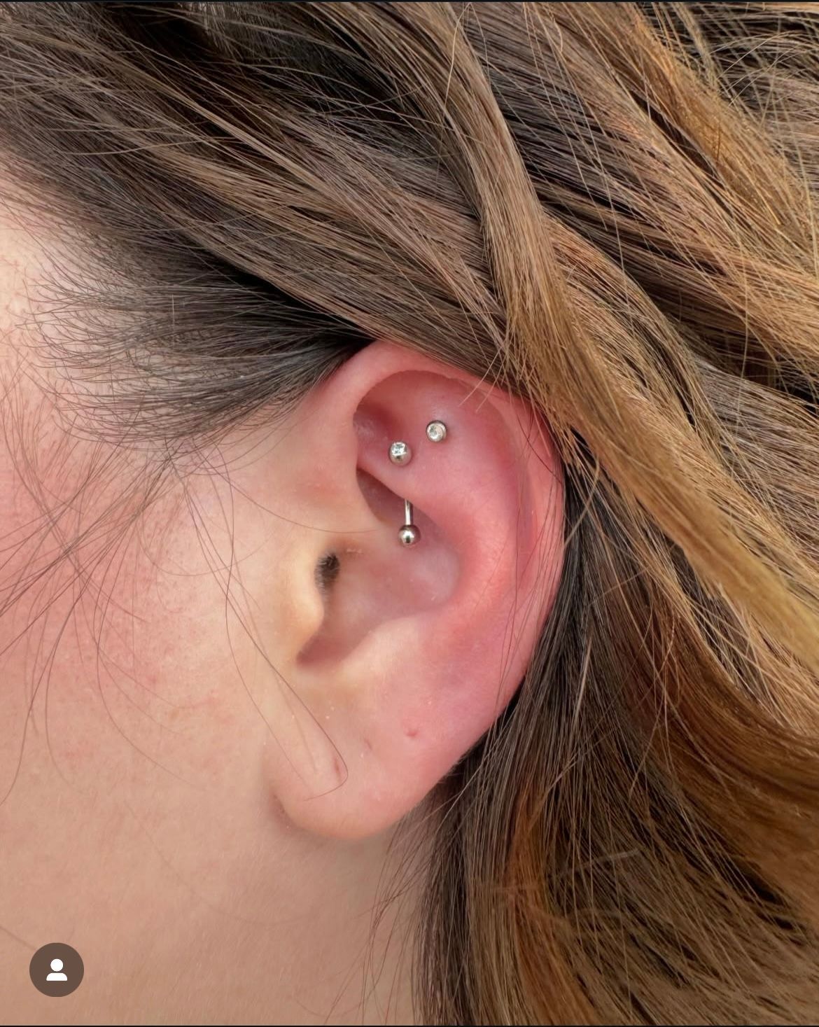 Close-up of an ear with three piercings: one curved barbell and two small studs, against wavy brown hair.