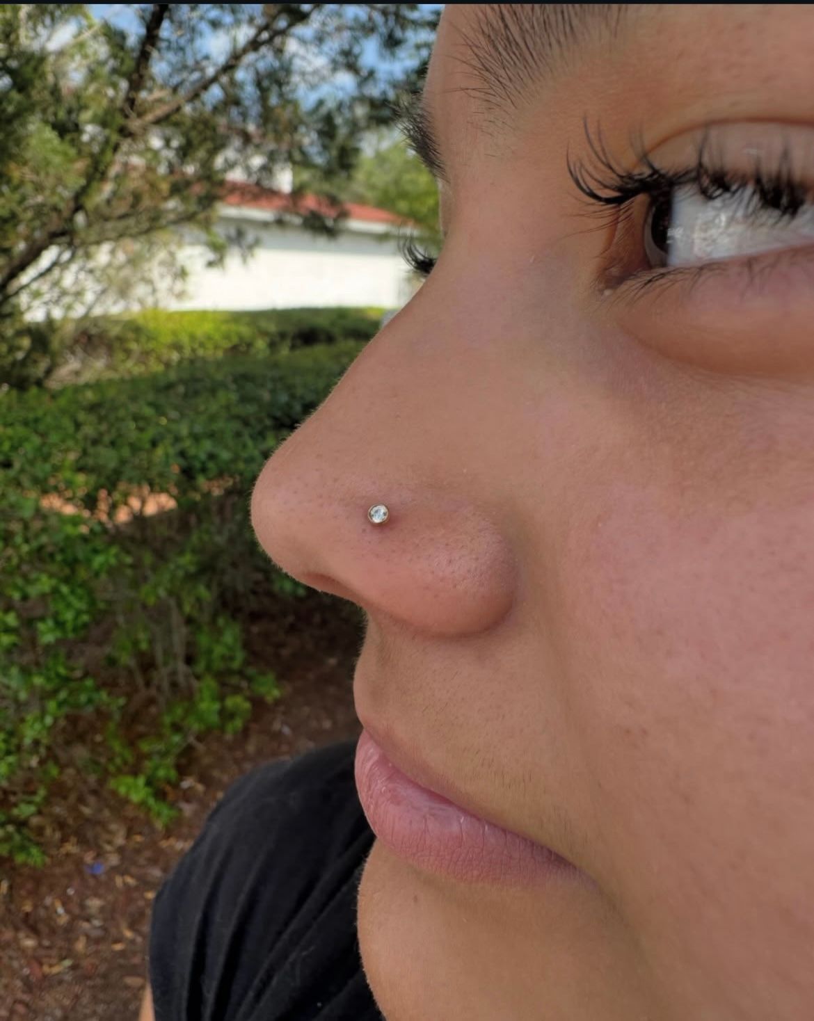 Person with a nose piercing; outdoors with greenery. A small gem is in the nostril.