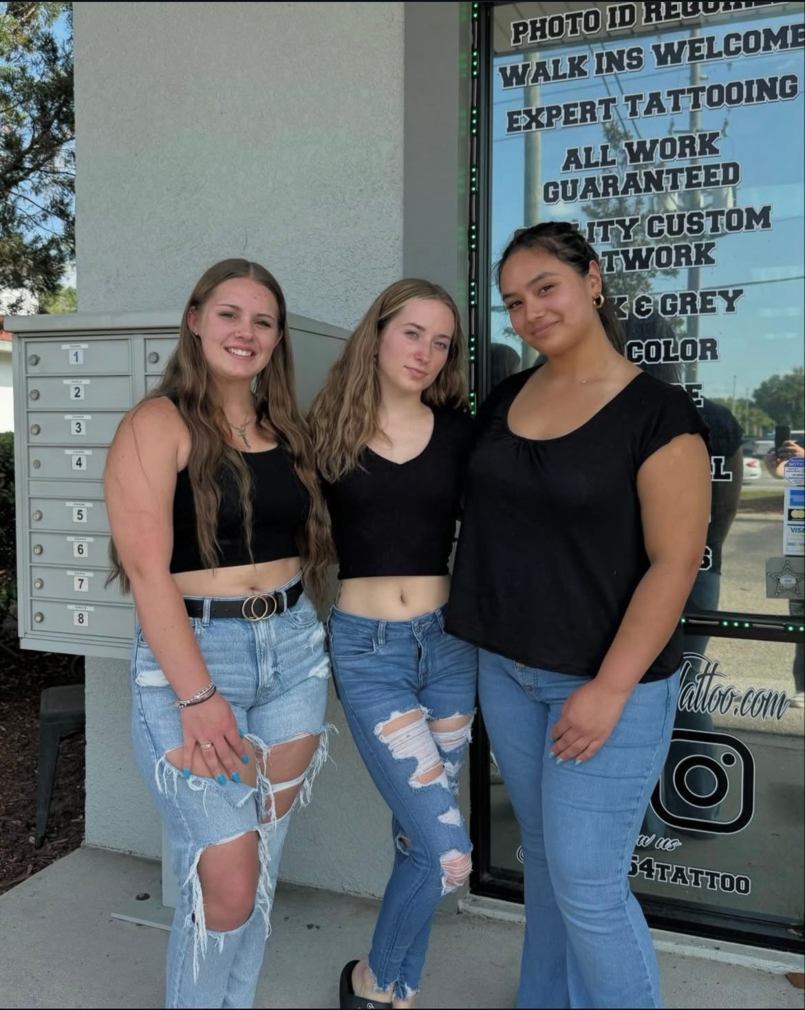 Three young women posing outside a tattoo parlor; all wearing black tops and jeans.