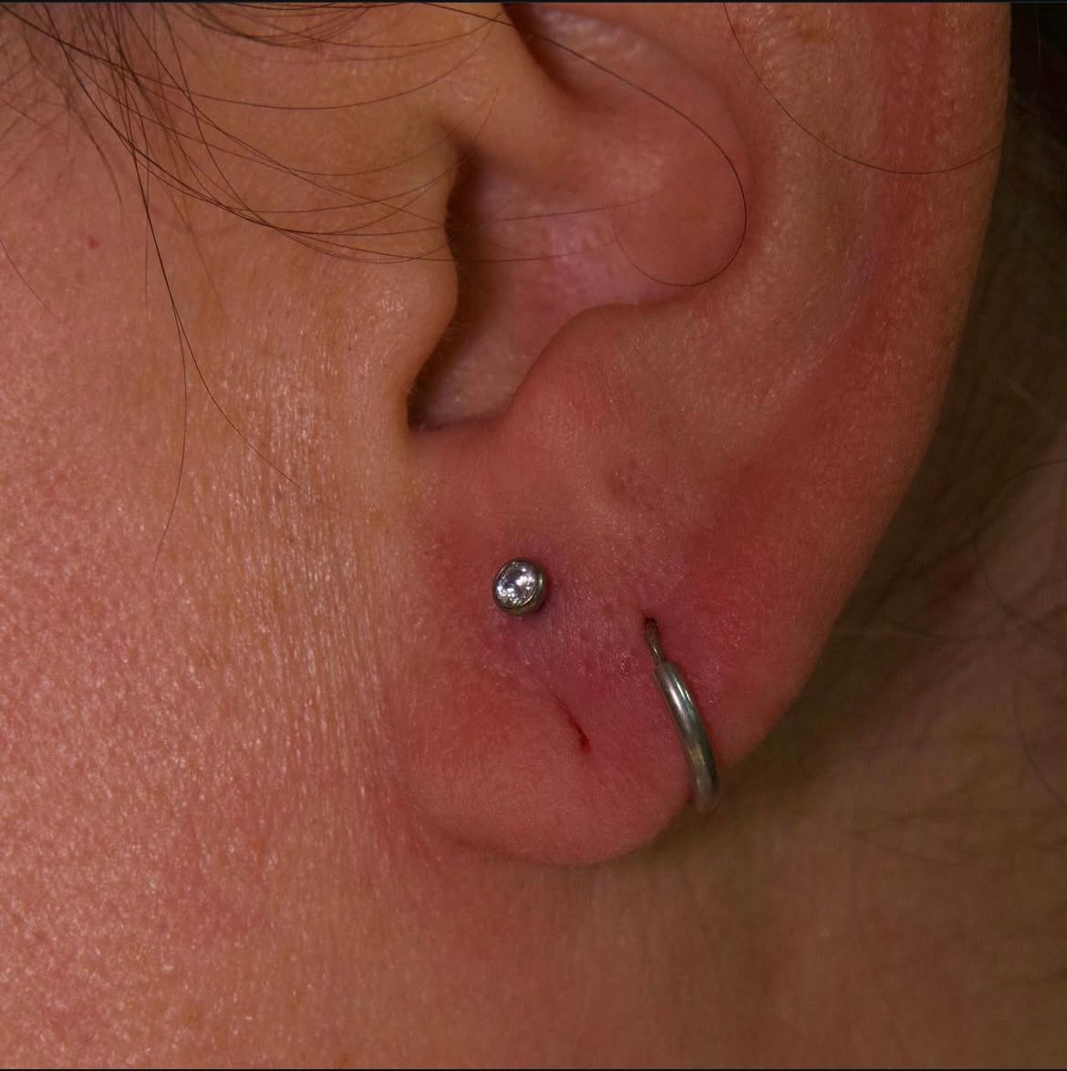 Close-up of a person's ear with two piercings: a stud and a hoop earring. The skin is light-toned.