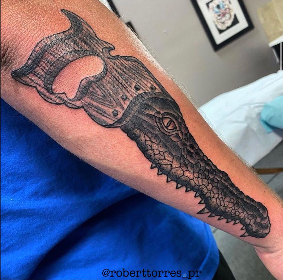 Tattoo of a crocodile's head with saw teeth, integrated with the handle of a handsaw on a forearm.