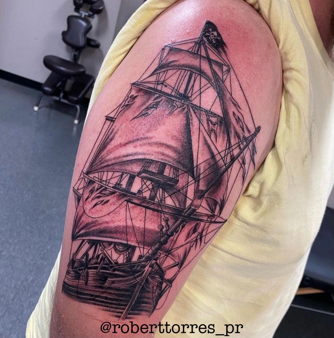 Black and white pirate ship tattoo on a person's upper arm with a skull and crossbones flag.