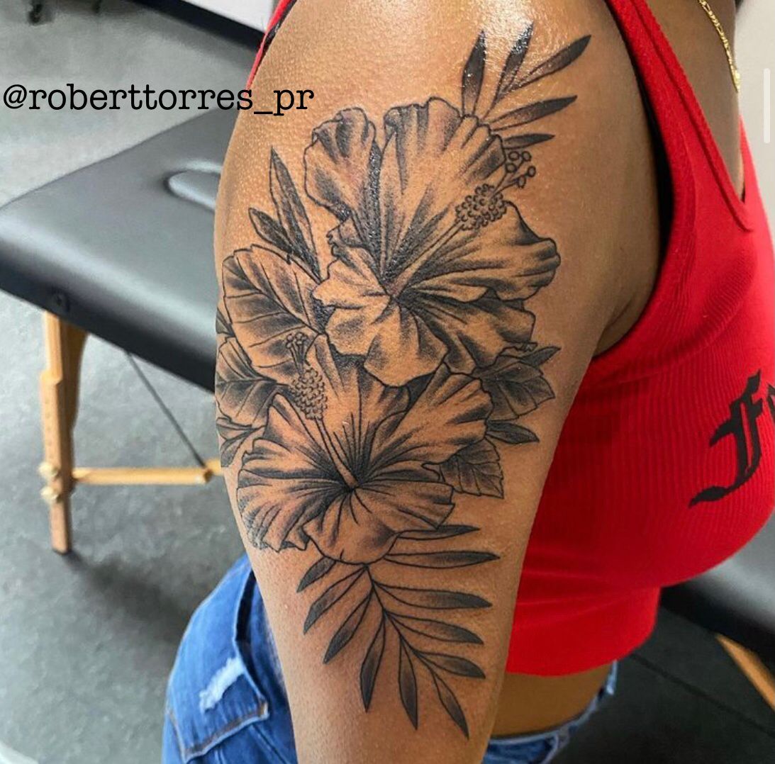 Tattoo of hibiscus flowers and leaves on a woman's arm, black ink. She's wearing a red top.