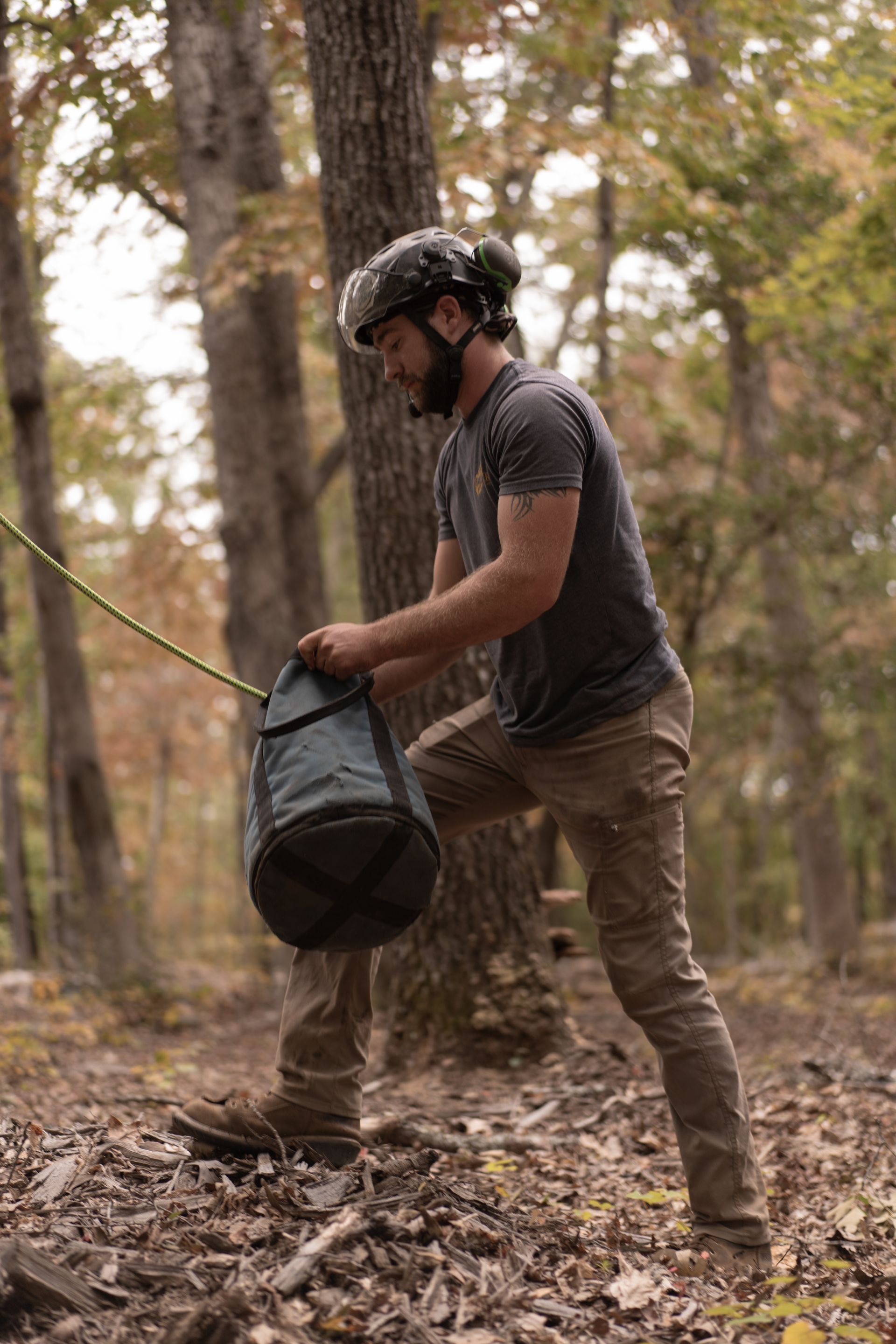 Person in a helmet filling a hanging black bag in a wooded outdoor area.