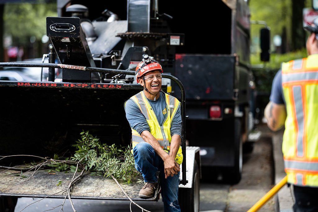 Worker in orange safety vest resting beside a roadwork machine on a street, holding a shovel.