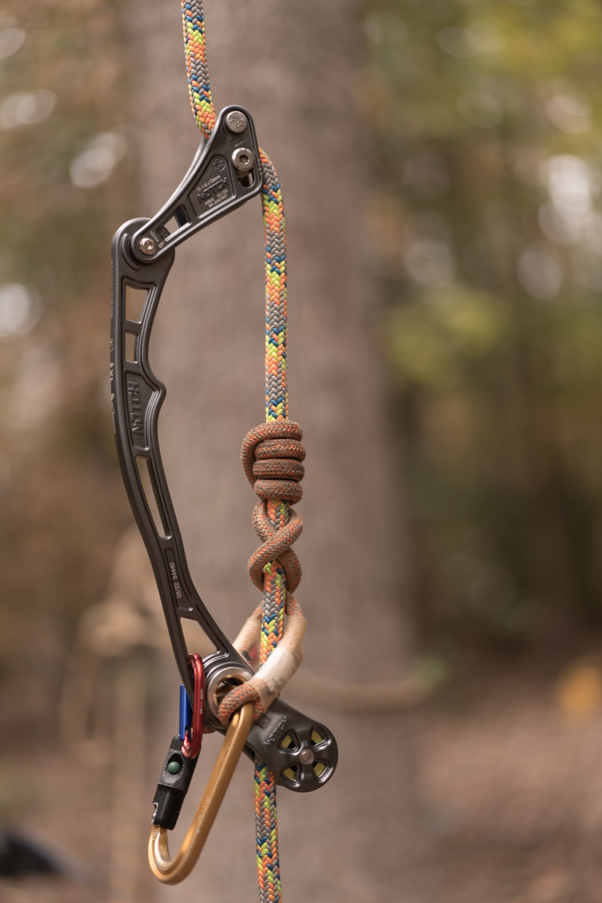 Black climbing belay device clipped to a multicolored rope, with a knot against a blurred outdoor background