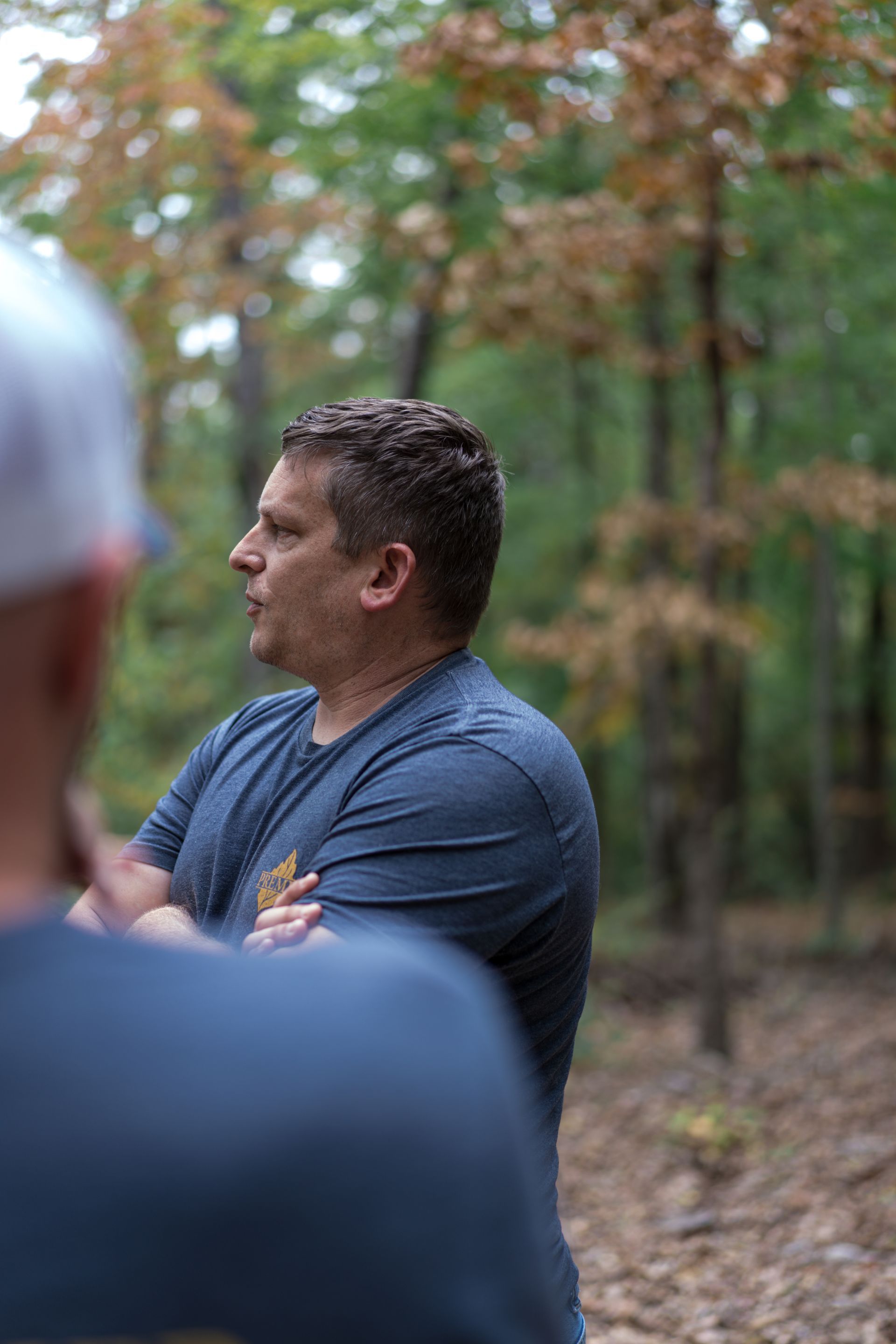 Man in a blue shirt standing with arms crossed in a wooded outdoor setting