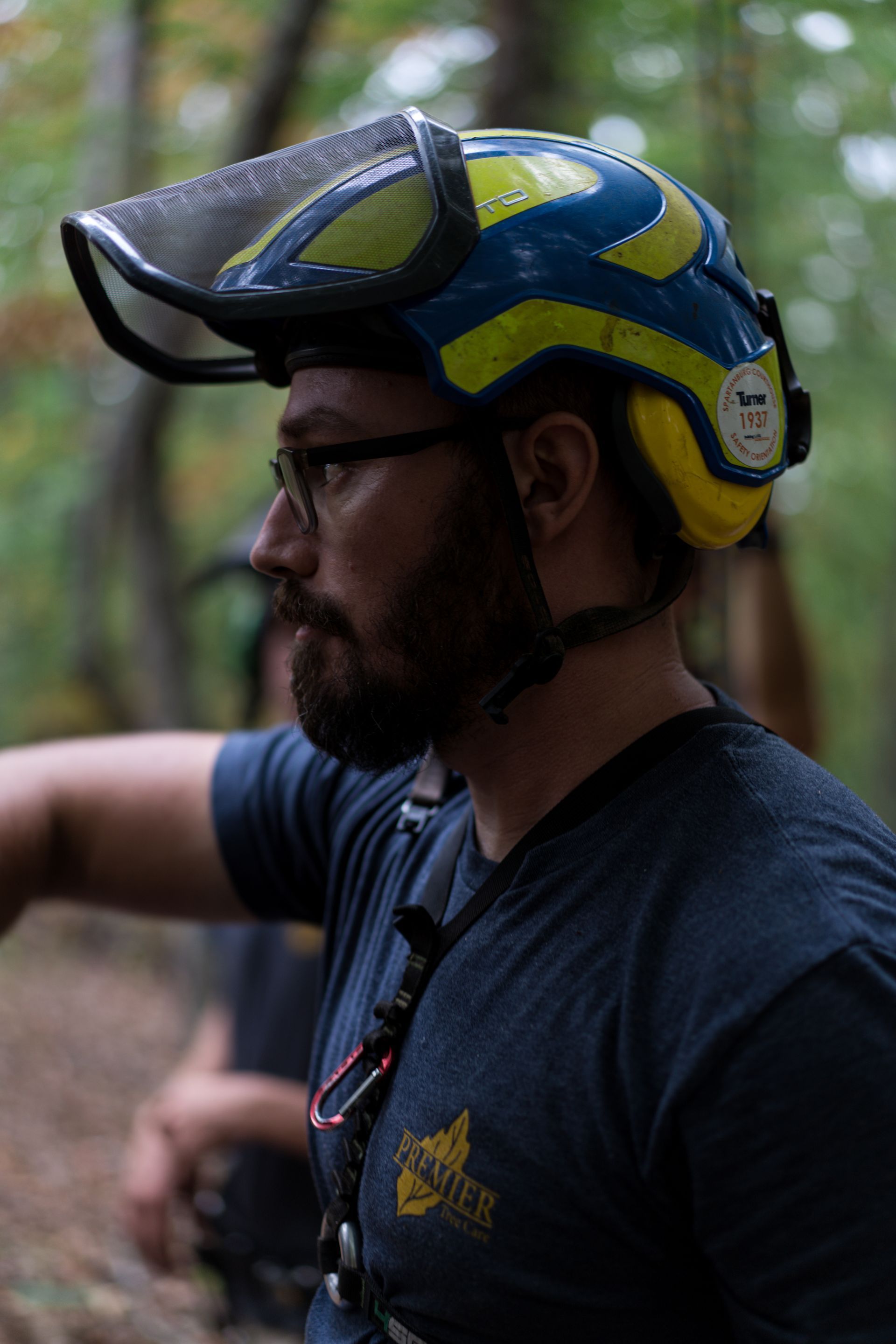 Side-profile of a man wearing a blue-and-yellow helmet and glasses in a wooded outdoor setting