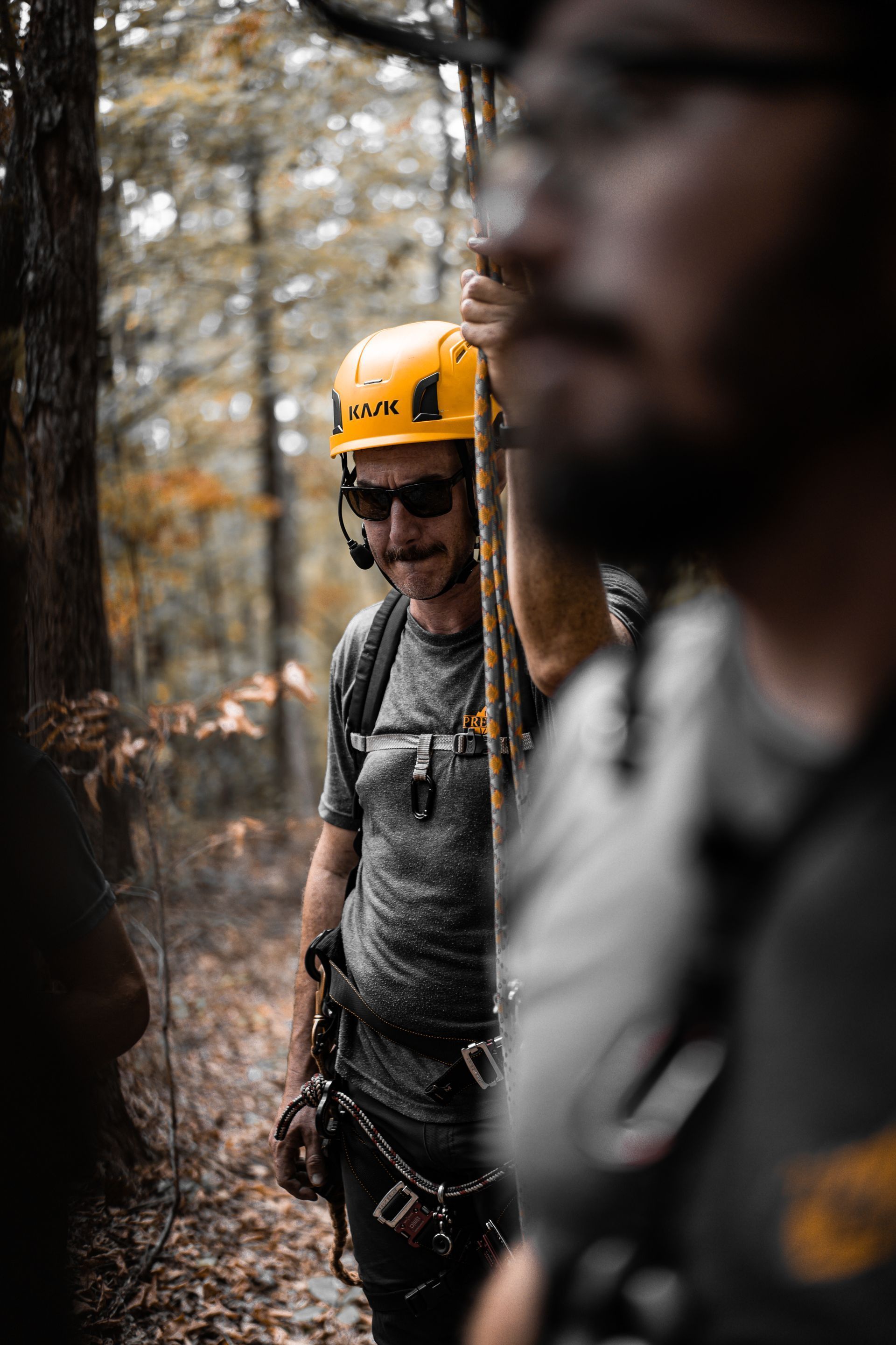 Two climbers in helmets on a forest trail, one in focus with a yellow helmet and rope gear.