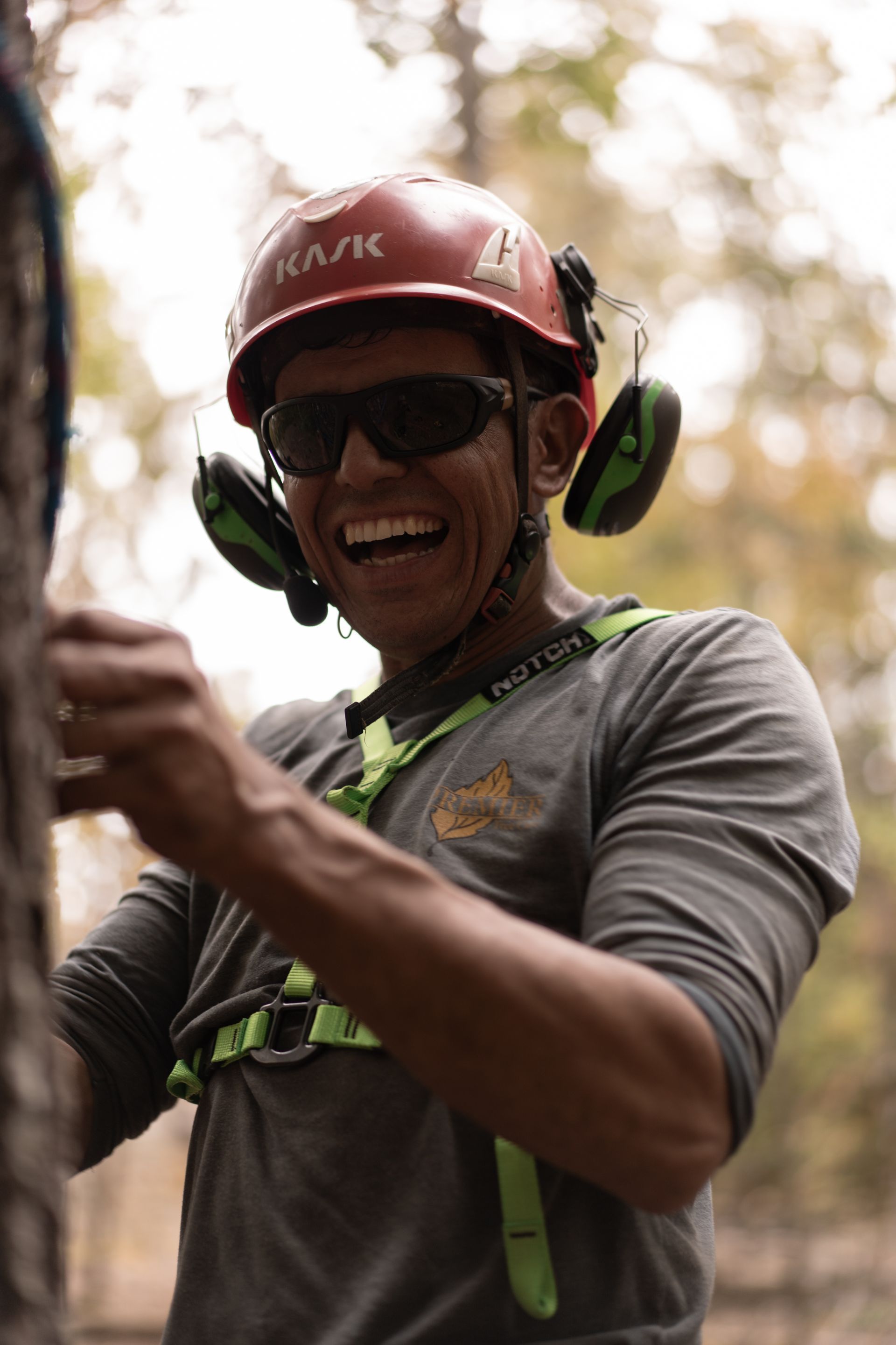 Smiling climber in a red helmet and sunglasses, wearing a gray shirt and green harness outdoors