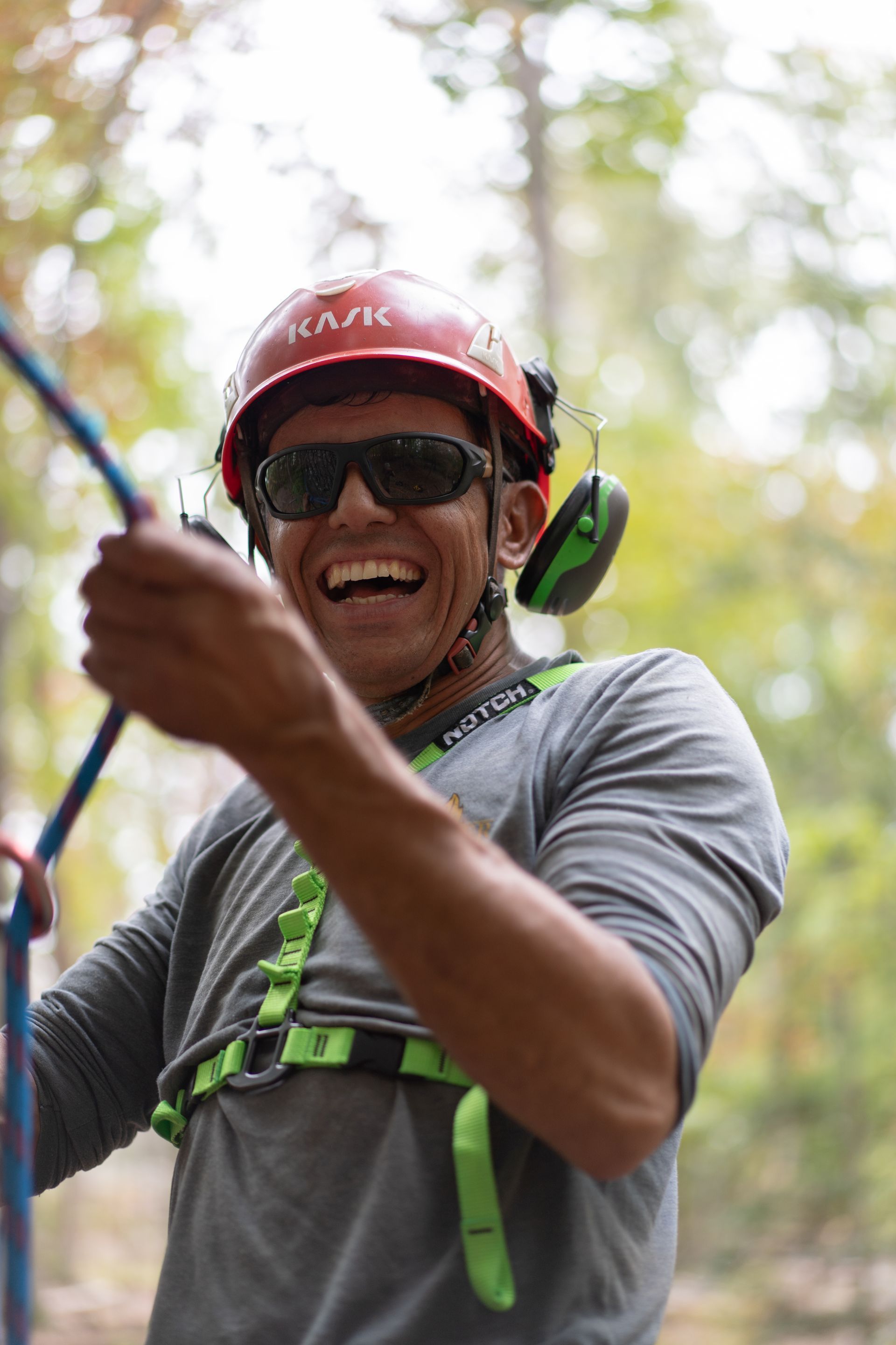 Smiling climber in red helmet and harness, reaching while ziplining or rope climbing in a wooded setting