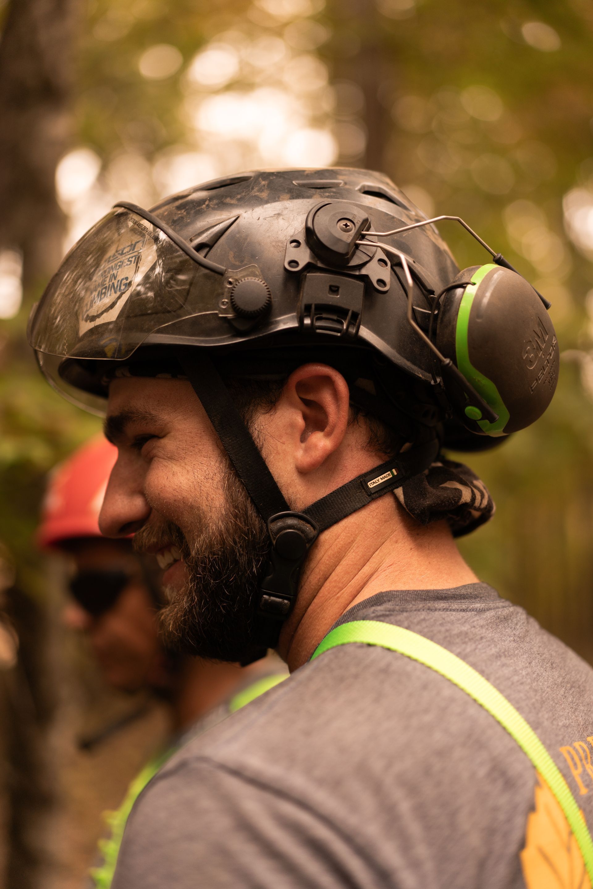 Helmeted worker in profile wearing a gray shirt and reflective straps in a wooded outdoor setting