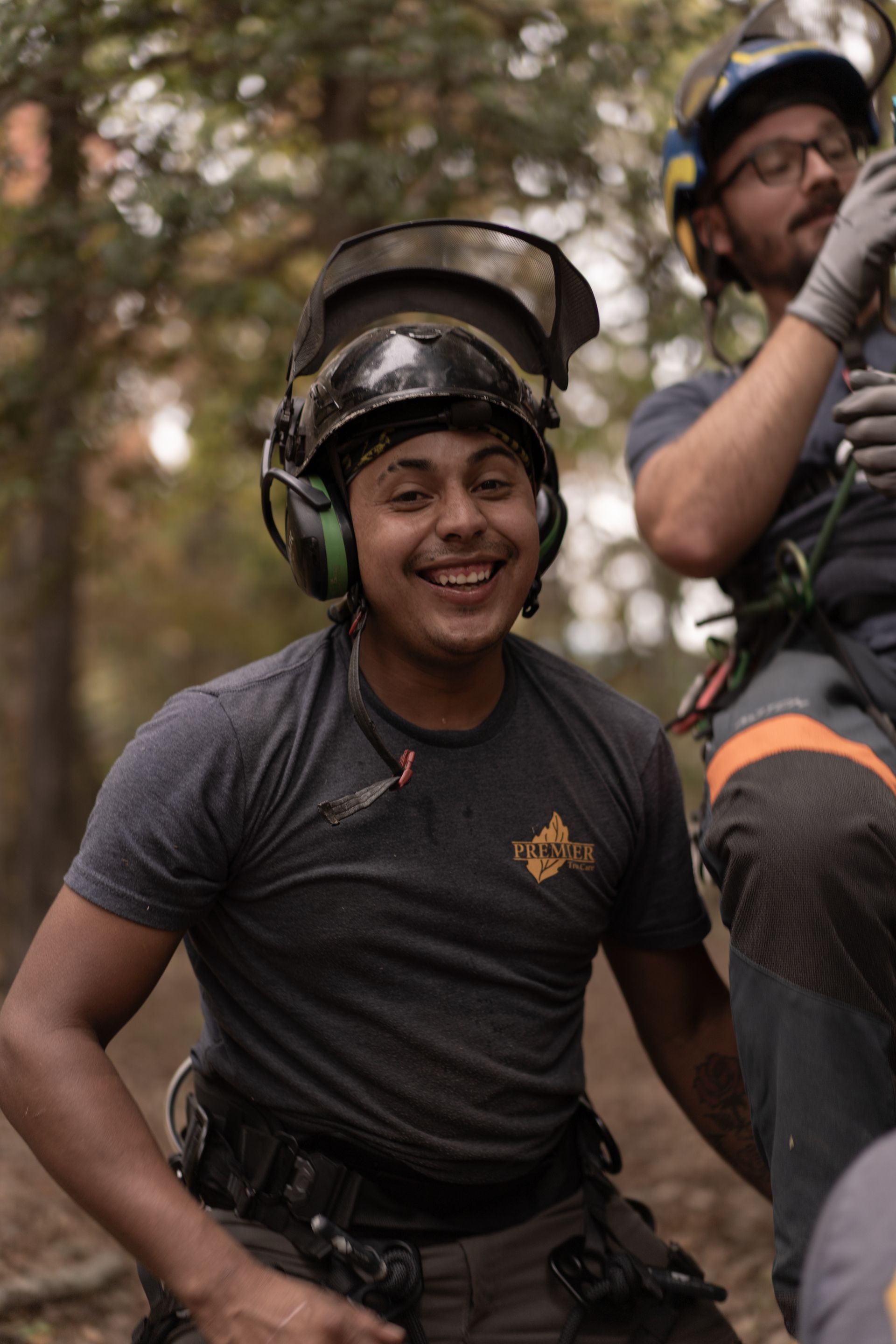 Smiling climber in a black helmet and gray shirt with a harness, outdoors among trees