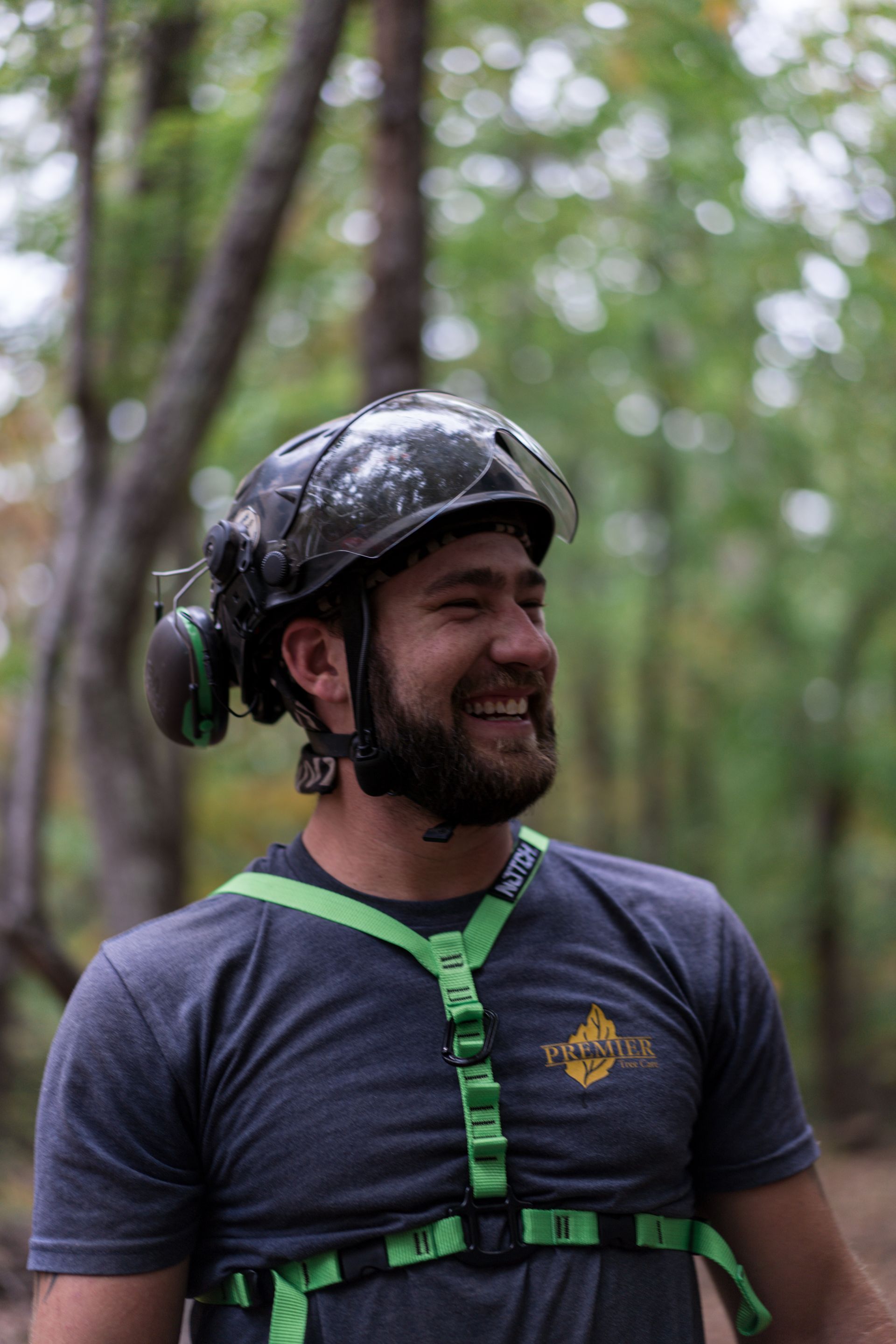 Smiling helmeted climber in a forest, wearing a gray shirt and green harness