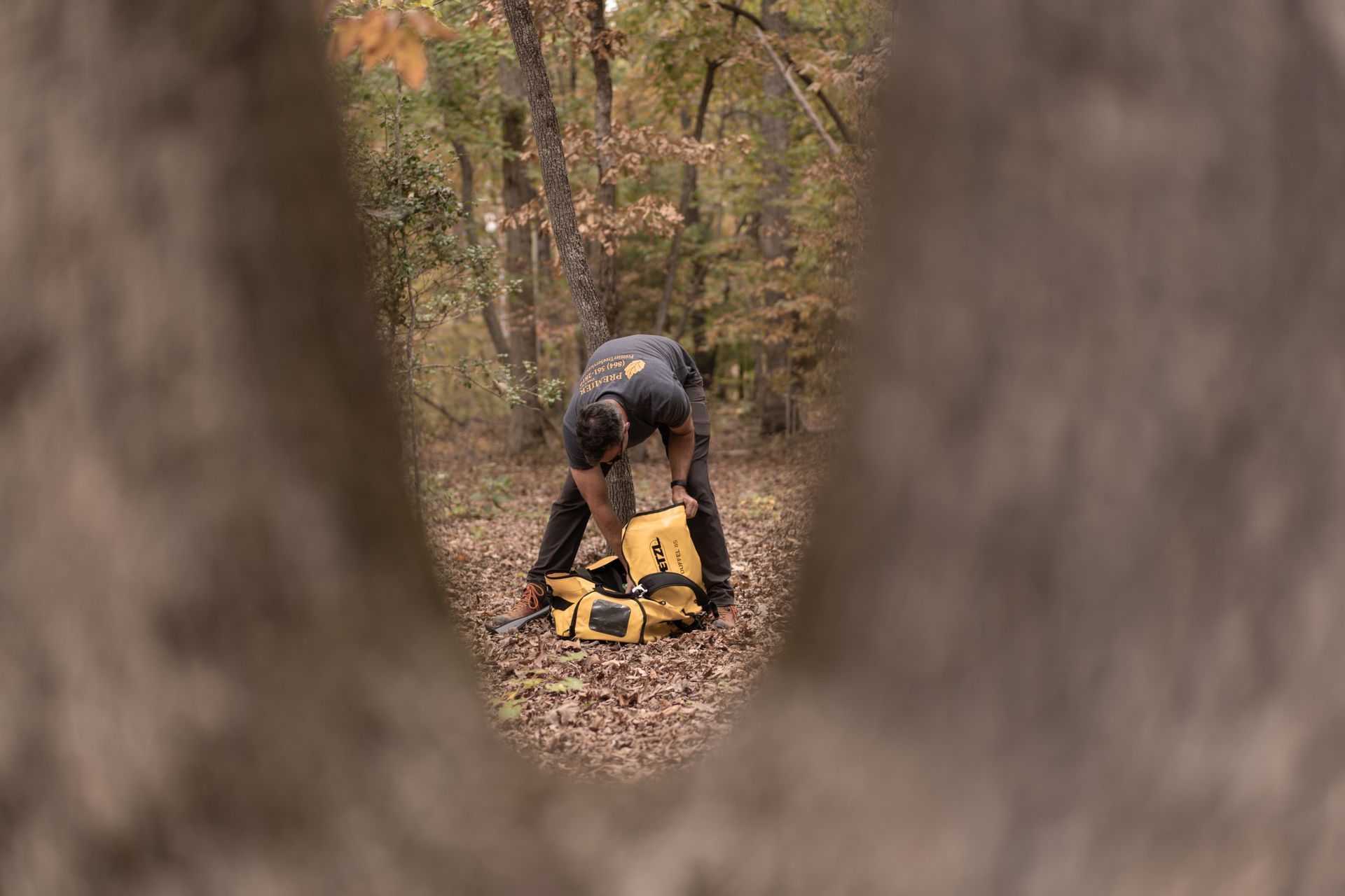 Person bending over a yellow backpack in a wooded path, framed between tree trunks