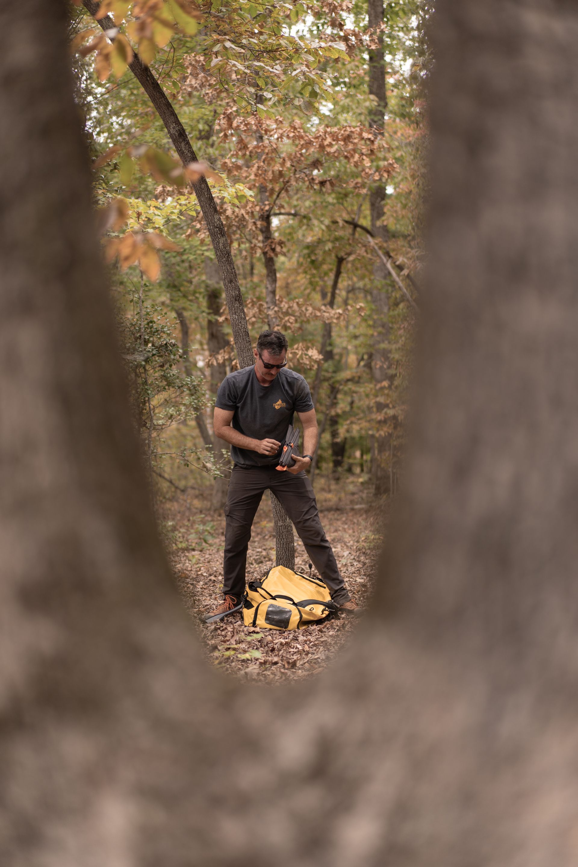 Man standing in a forest, holding a tool beside a yellow backpack, framed by tree trunks.