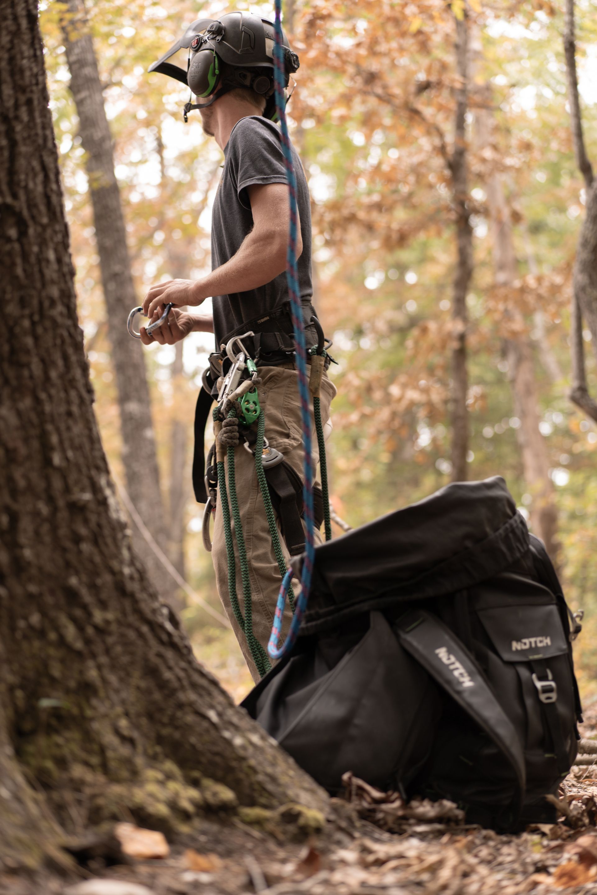 Person in a helmet standing by a tree with climbing gear and a backpack in an autumn forest
