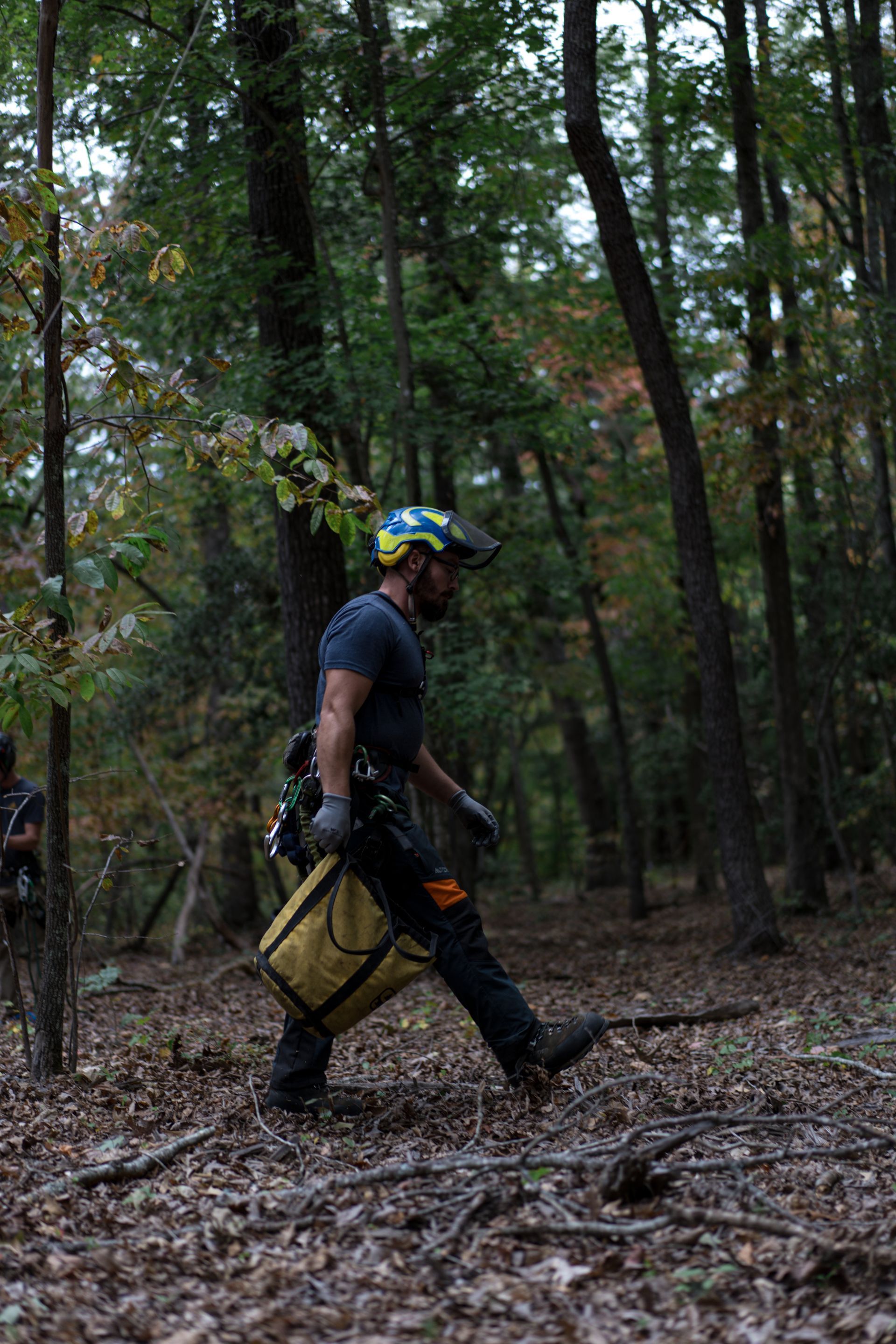 Person in a blue helmet and dark clothes walking through a wooded forest carrying gear and a yellow bag