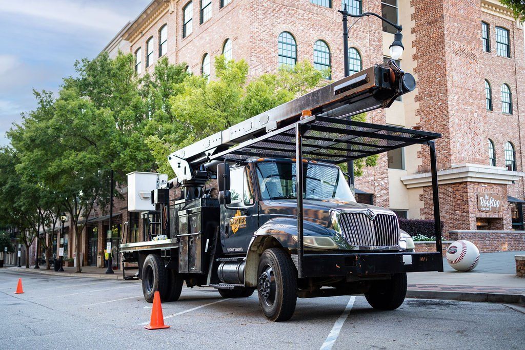 Utility truck with a boom lift parked on a tree-lined city street beside a brick building.