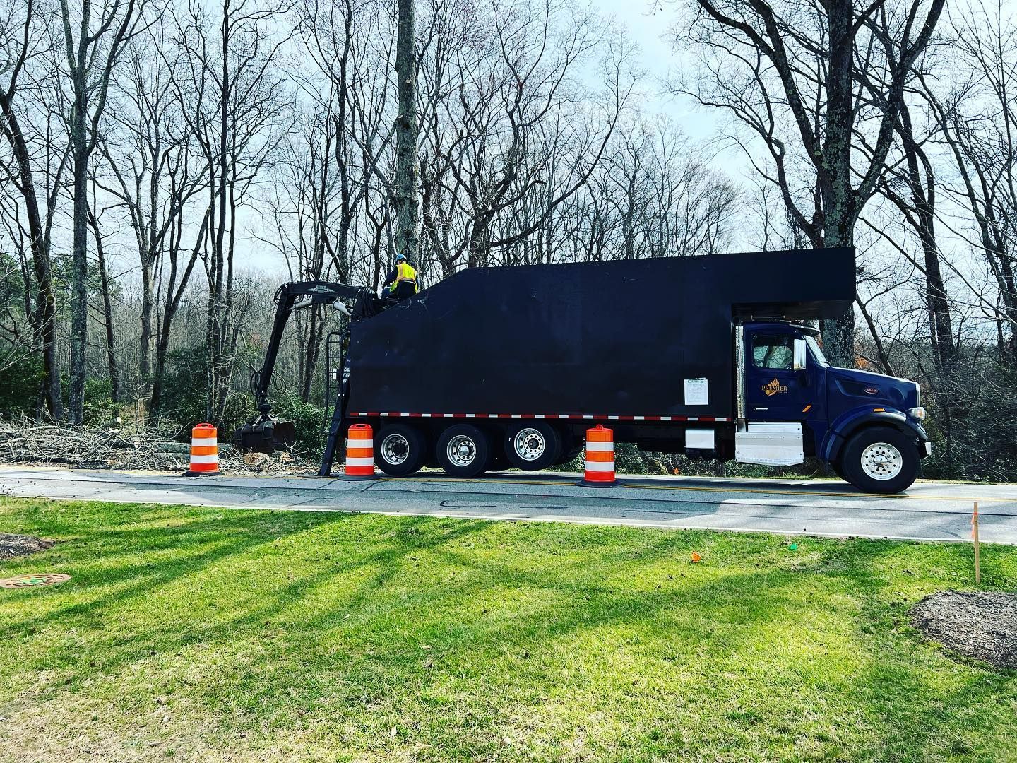 Blue utility truck parked on a road beside a grassy lawn, with orange traffic cones and leafless trees nearby